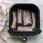 raw pork bacon laying in an air fryer basket with a red and white towel to the left of the basket