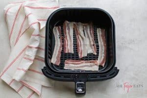 raw pork bacon laying in an air fryer basket with a red and white towel to the left of the basket
