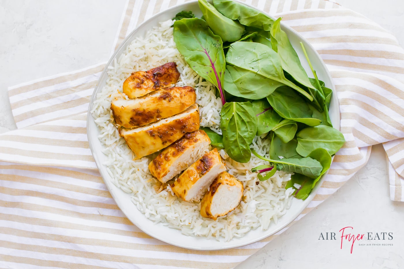 A sliced browned chicken breast arranged over a plate of white rice aside a pile of fresh spinach