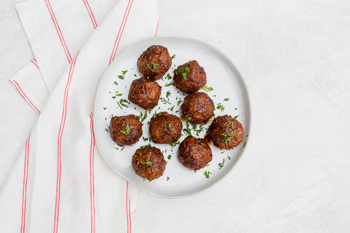 9 meatballs sprinkled with green parsley arranged on a white plate aside a red and white striped towel