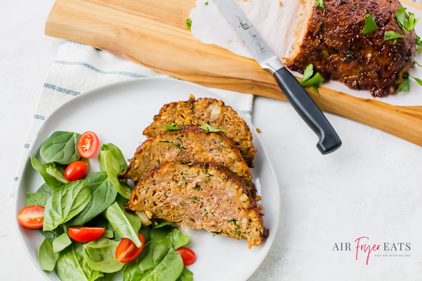 slices of meatloaf on a white plate with a spinach salad aside a cutting board with the remaining meatloaf and a knife