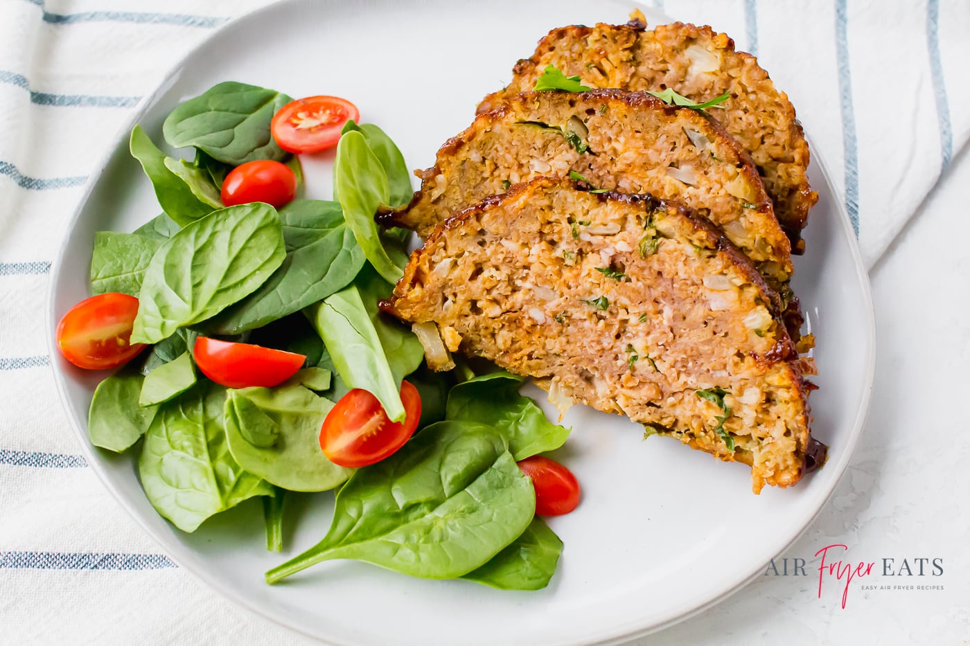 three slices of glazed meatloaf topped with parsley and served with a spinach and tomato salad on a round white plate