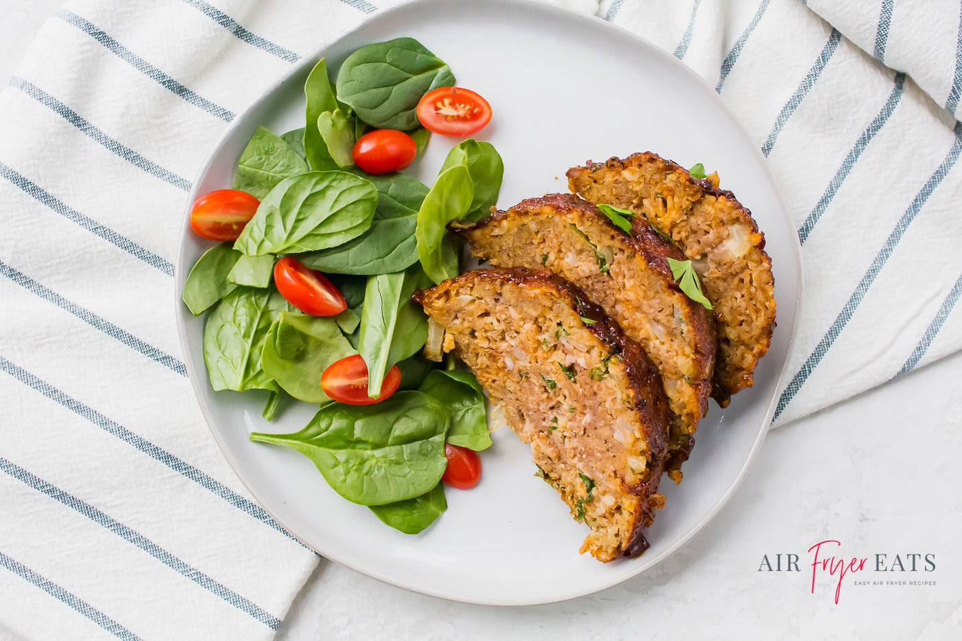 slices of meatloaf topped with herbs and arranged beside a spinach and tomato salad on a white plate