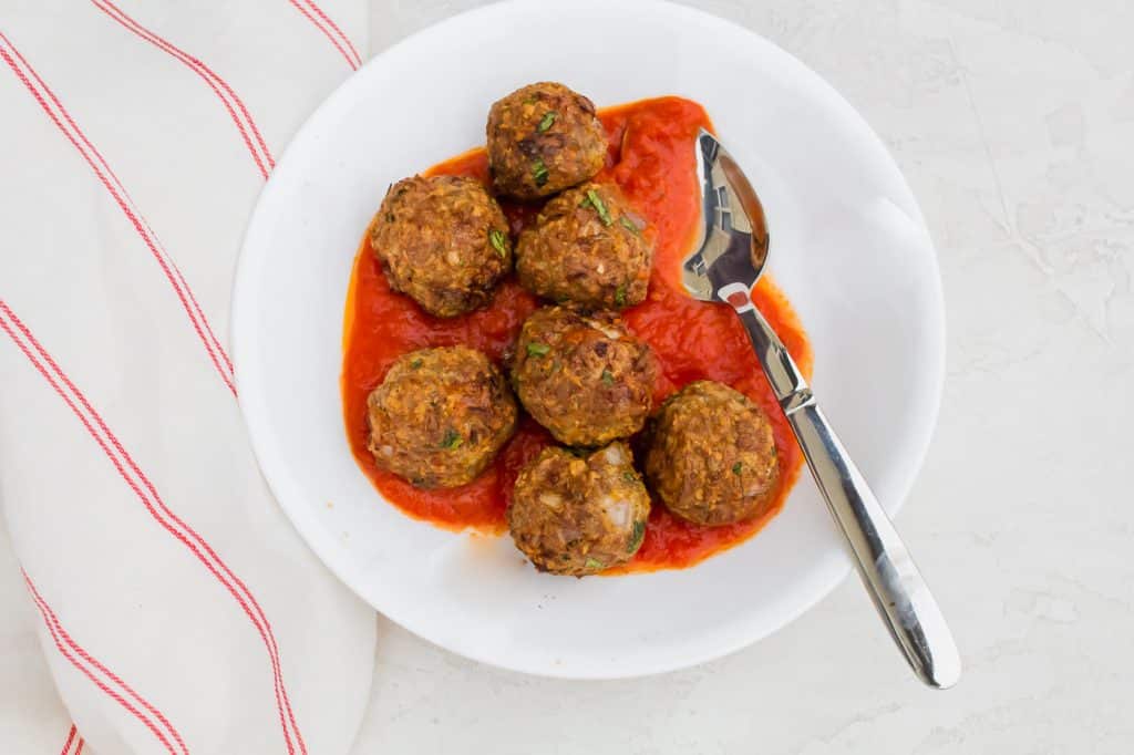 seven brown meatballs over red tomato sauce in a white bowl with a silver spoon