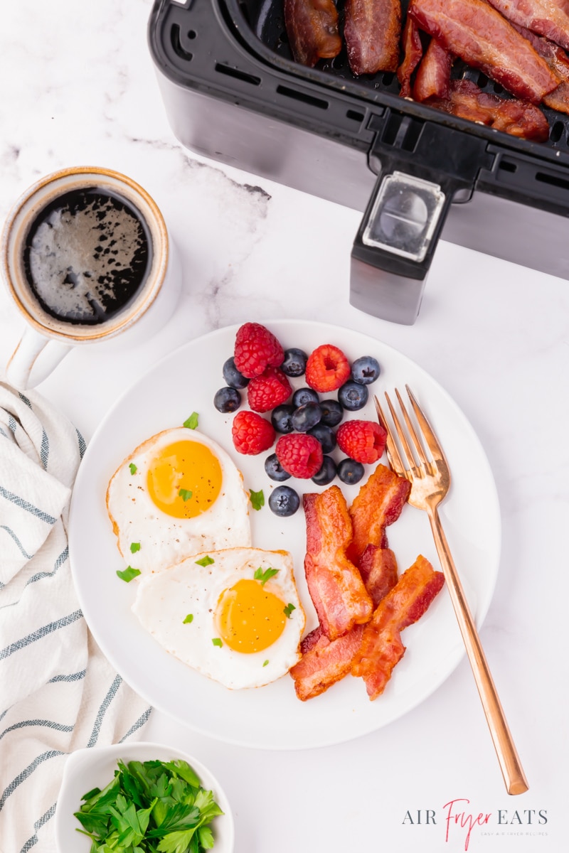 a breakfast plate of two sunny side up eggs, strips of bacon, and fresh raspberries and blueberries. A gold fork rests on the plate, a cup of coffee is above it. The air fryer is off to the side.