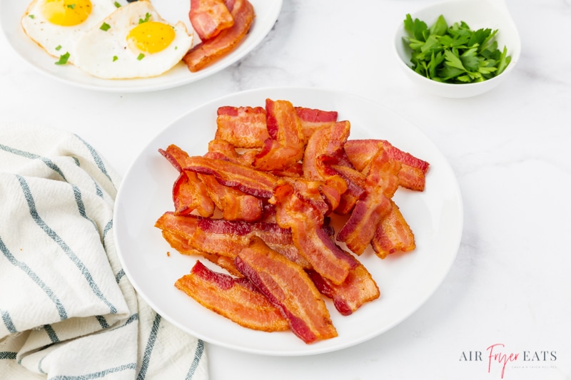a white plate of air fried bacon on the breakfast table next to a plate of eggs