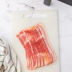 a slab of raw sliced bacon on a plastic cutting board in front of a cosori air fryer