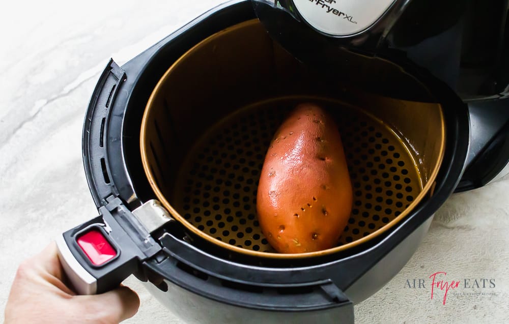 A hand placing an air fryer basket holding a large sweet potato into an air fryer