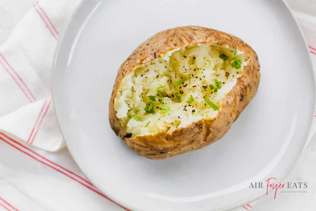 a baked potato split with black pepper and green herbs on a white plate