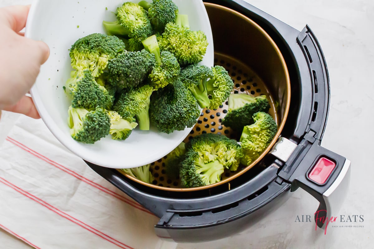 a hand pouring broccoli florets from a white bowl into the basket of an air fryer