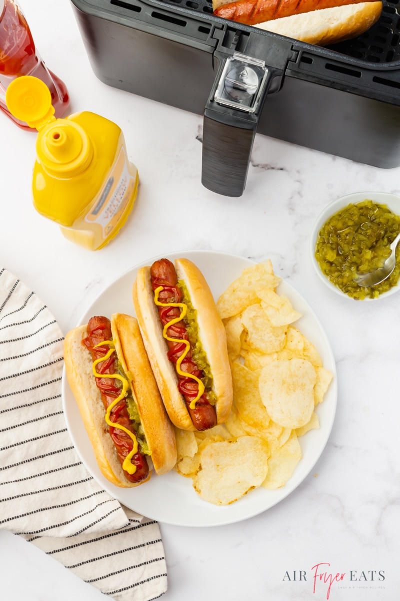 a plate of to hotdogs in rolls, topped with relish, ketchup and mustard, with a side of potato chips. The condiment bottles are above the plate, and a cosori air fryer is also there. Viewed from above. 