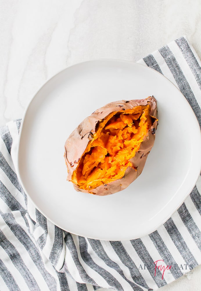 A baked sweet potato with crispy skin split open on a white round plate