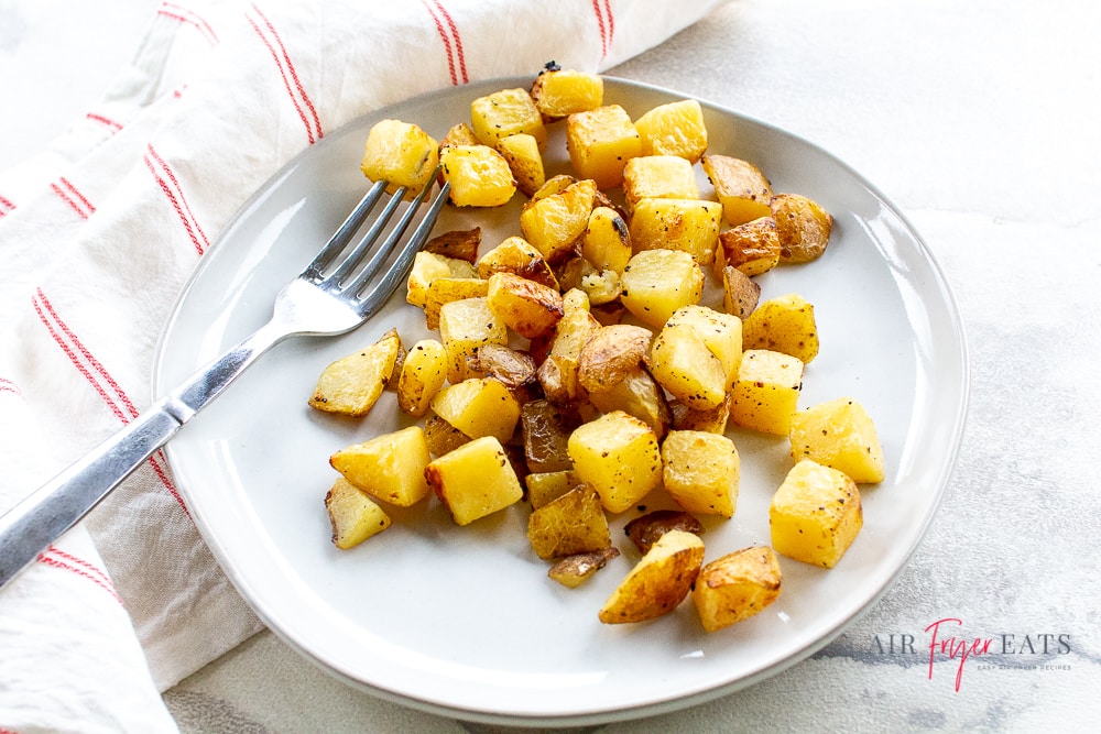 landscape style photograph of white plate with golden brown potatoes cubes on it along with a silver fork.