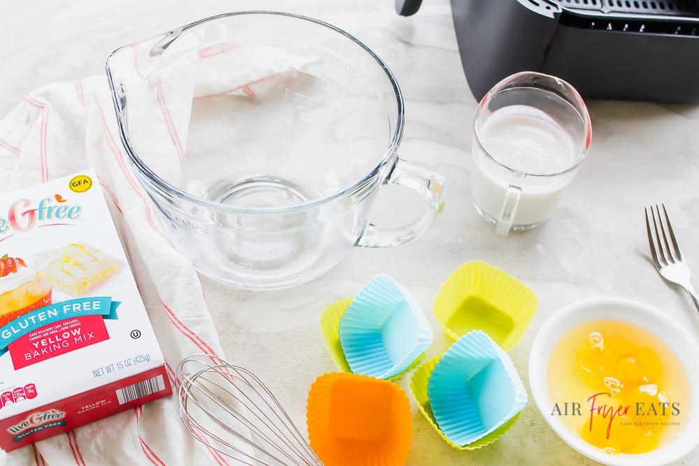 whisk, measuring cups, fork, white bowl with raw eggs, box mix of cake mix and black air fryer basket, all pictures