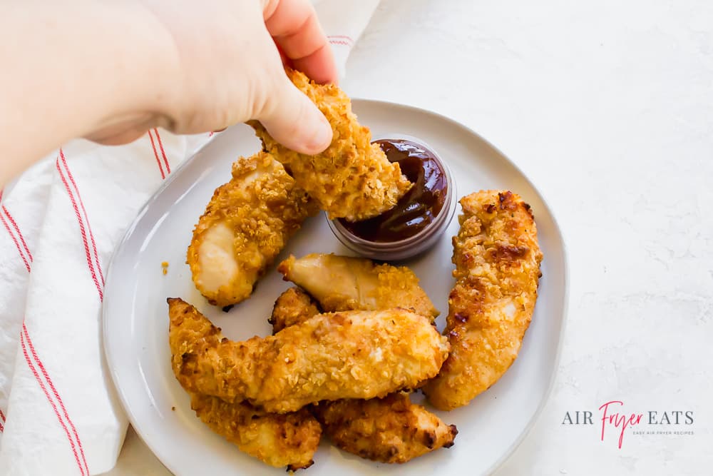 a hand dipping an air fryer chicken tender into a small cup of barbecue sauce