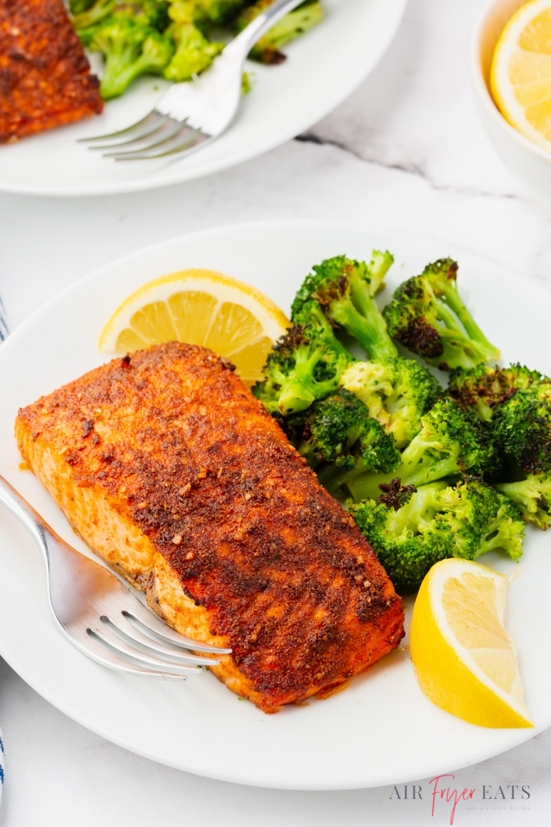 A white plate with a browned seasoned salmon filet and a side of air fryer broccoli, garnished with lemon wedges.