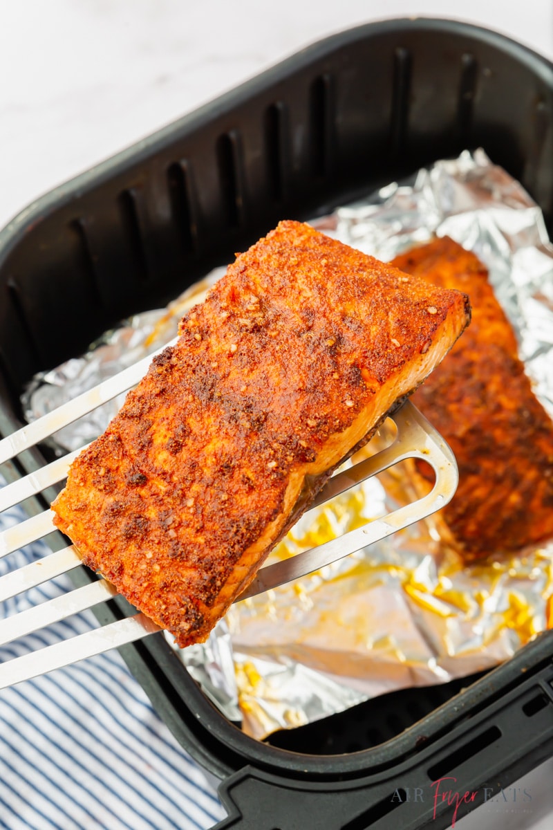 a piece of salmon being lifted out of an air fryer basket lined with foil. 