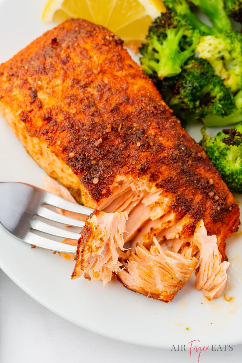 a piece of air fryer salmon on a plate with broccoli and lemon wedges. A fork is flaking a bite off. 
