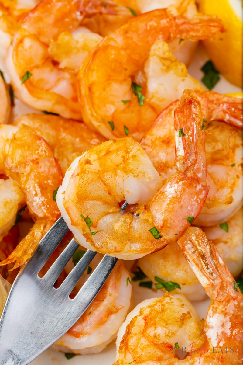 A zoomed in close up photo of a pile of cooked shrimp. Shrimp is golden in color with hues of orange and some red on the tails.  The head of a silver fork is poked through one of the shrimp. Small pieces of diced green herbs are scattered on top of the shrimp.