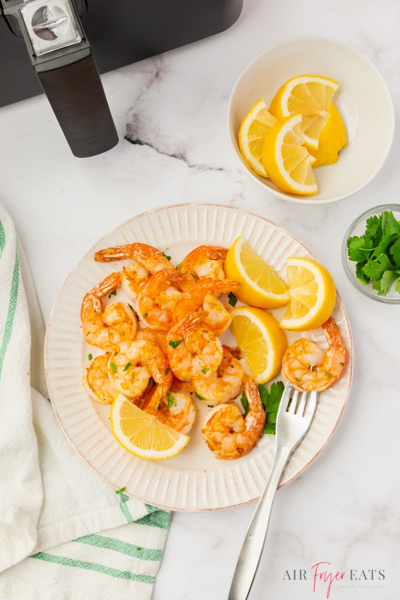 a plate of cooked shrimp garnished with parsley leaves and lemon wedges sits on top of a green and white striped kitchen towel. Behind the plate is a cosori aif fryer, and to the right is a bowl of lemon wedges and a bowl of parsley