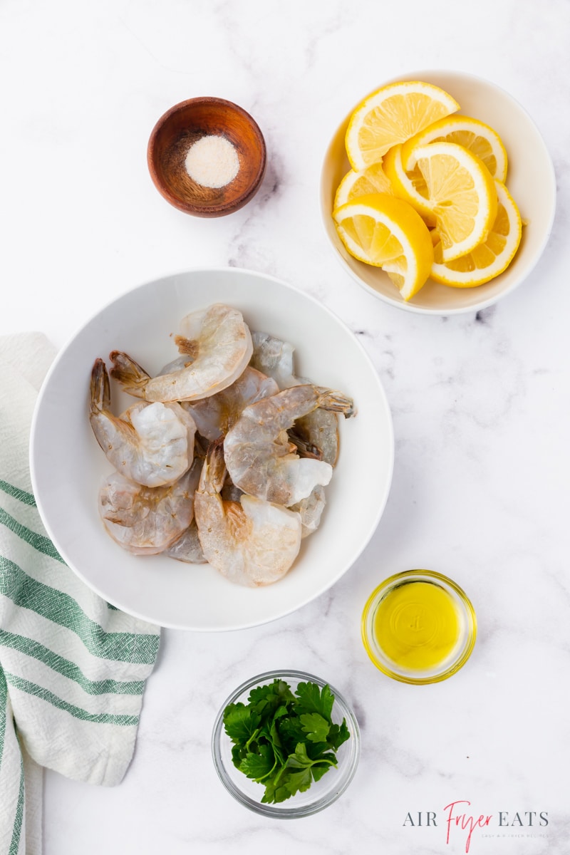 The ingredients needed to make shrimp in the air fryer, including raw shrimp, oil, and lemons, each in a separate bowl on a marble countertop