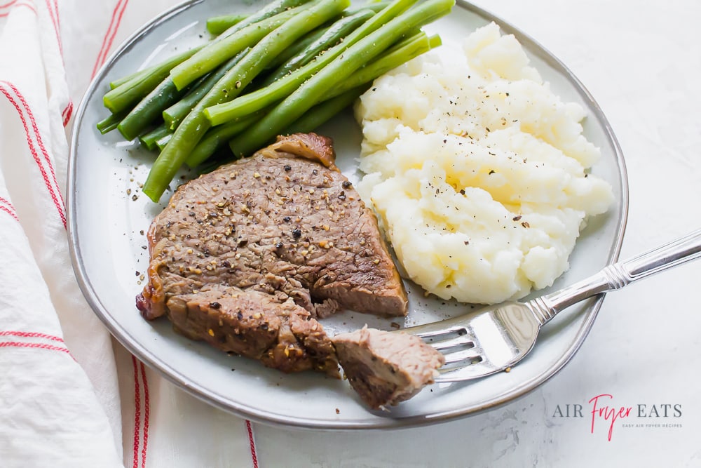 a fork piercing a piece of steak from a steak filet on a plate with mashed potatoes and green beans