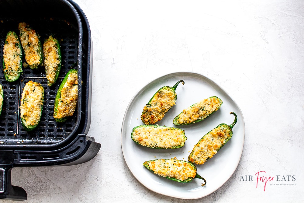 White plate with 5 air fried jalapeno poppers. To the left of the plate is a black air fryer basket with 5 cooked air fryer jalapeno poppers. This is set on a white back ground.