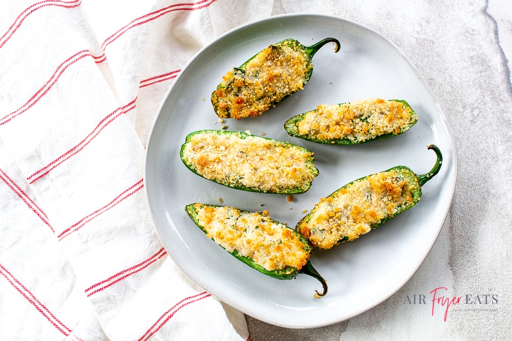 five air fryer jalapeno poppers cooked and on a white plate with a white napkin with red stripes to the left. All on a white background.