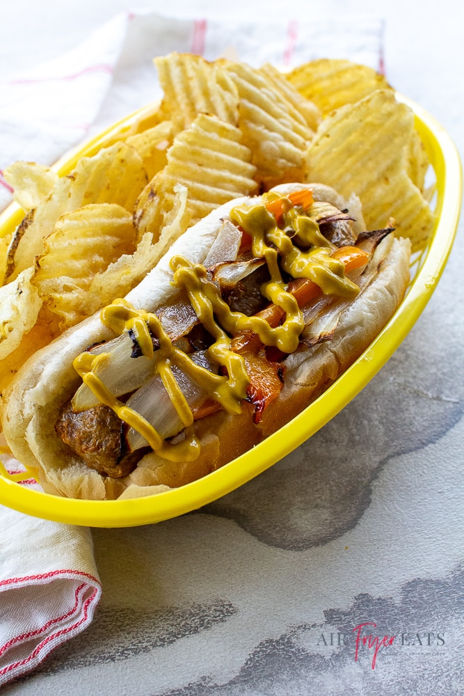 brats, bell peppers, and onions on a hot dog bun with chips on a yellow plate