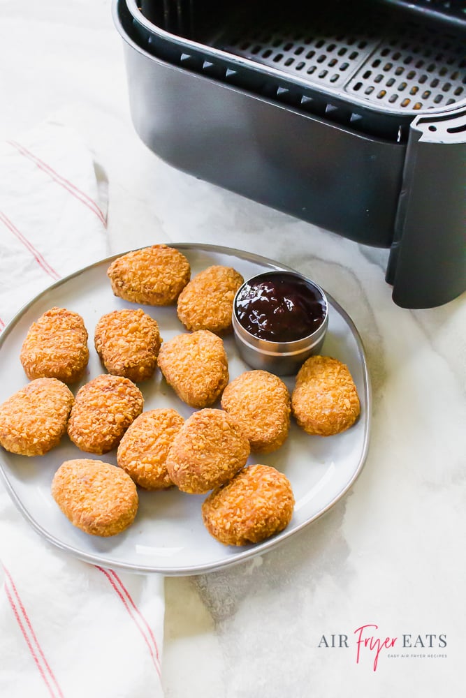 Vertical picture with black air fryer basket in the top right and golden brown cooked nuggets on a white plate with red dipping sauce in the bottom portion of the picture.