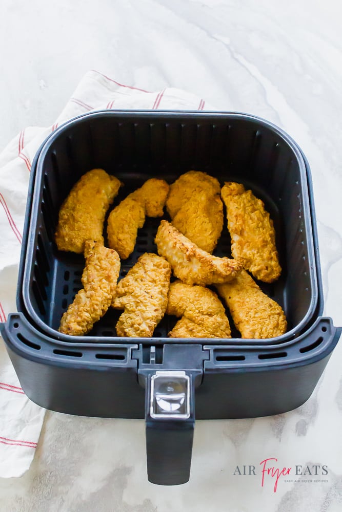frozen chicken strips in black air fryer basket on top of a white background with white napkin with thin red stripe