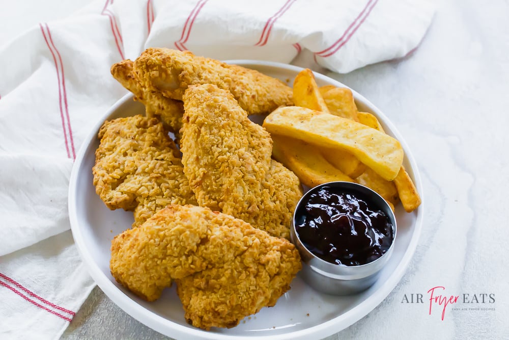 Air fried chicken tenders on a white plate with steak fries. BBQ sauce dipping bowl on a white background. White napkin with thin red stripe to the left and top of plate.