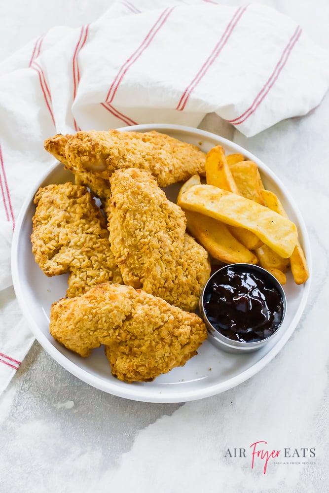 White plate full of air fryer chicken strips with air fryer steak fries and a BBQ dipping sauce. White napkin with thin red stripes on left side and top. White background.