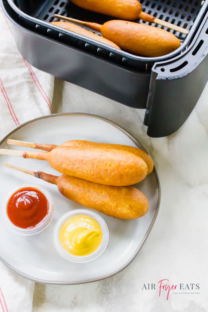 Vertical picture. Cooked breaded dogs in the air fryer basket in the top. Below that on an off white plate are breaded dogs with a cup of ketchup and mustard.