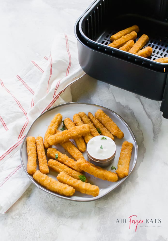 vertical picture of cooked fish sticks on white plate with white dipping sauce at the bottom of the picture. At the top of the picture is a black air fryer basket with fish sticks inside. To the left of the basket is a white napkin with thin red stripe.