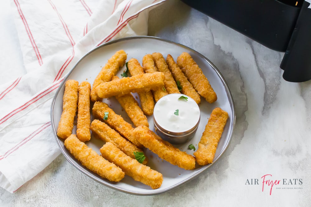 overhead angled shot of cooked sticks on a plate. They are golden brown on a white plate served with a white dipping sauce. there is a white napkin with thin red stripes to the left.