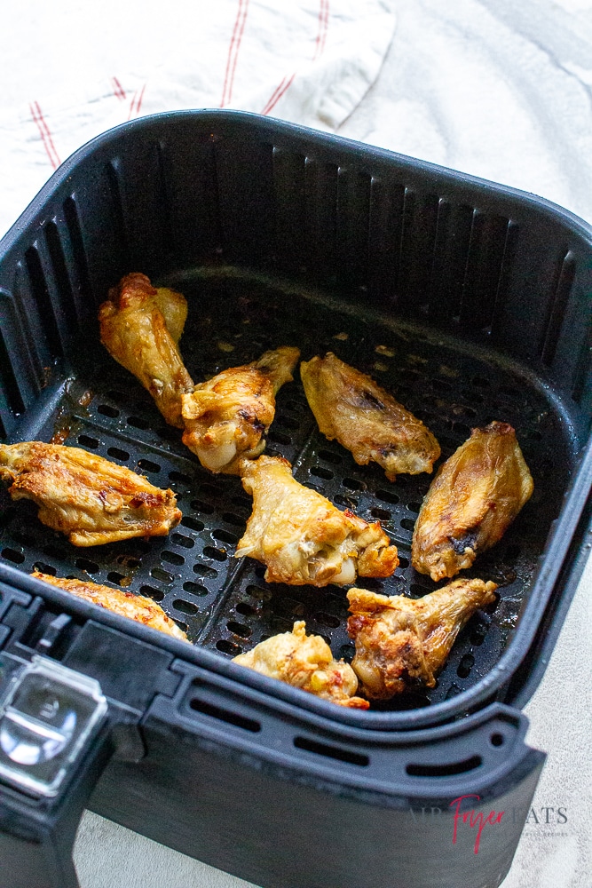 Vertical photo, showing a black air fryer basket with cooked chicken wings inside it. All on a white background with a white and red striped napkin to the left/top.