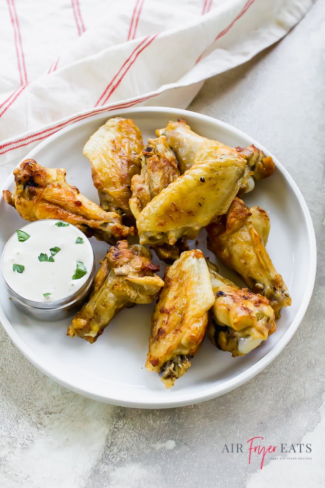 Air fryer frozen chicken wings, cooked and plated on a white plate with a cup of white ranch. The background is white and there is a white with a thin red stripe napkin to the upper left.