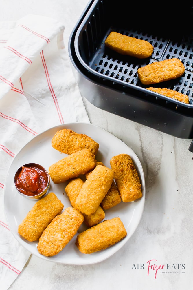 Vertical picture of cooked mozz sticks on a white plate with red dipping sauce. Black air fryer basket to the top right.