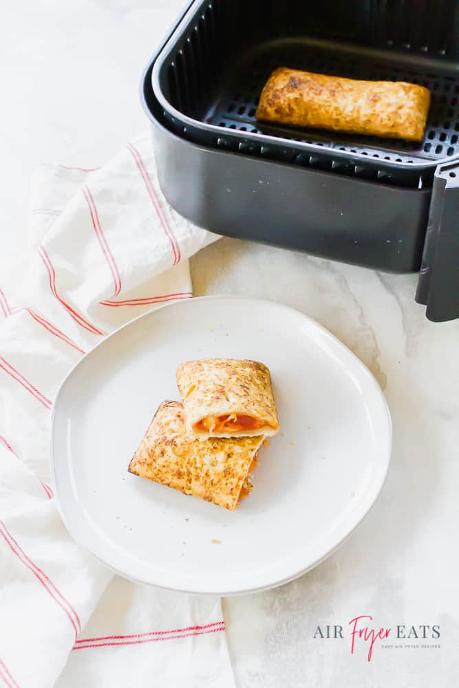 Vertical image. Cooked hot pocket in black air fryer basket at the top of the photo. At the bottom of the photo is an air fryer hot pocket sliced in half, stacked on a white plate.