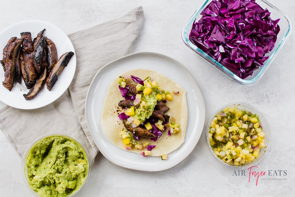 A mushroom taco surrounded by red cabbage, pineapple salsa, guacamole, and sliced portobello mushrooms