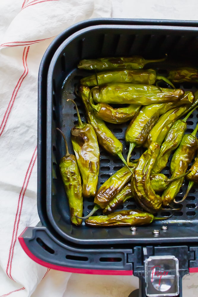 left side of a red and black air fryer basket. Inside the air fryer are blistered green shishito peppers.