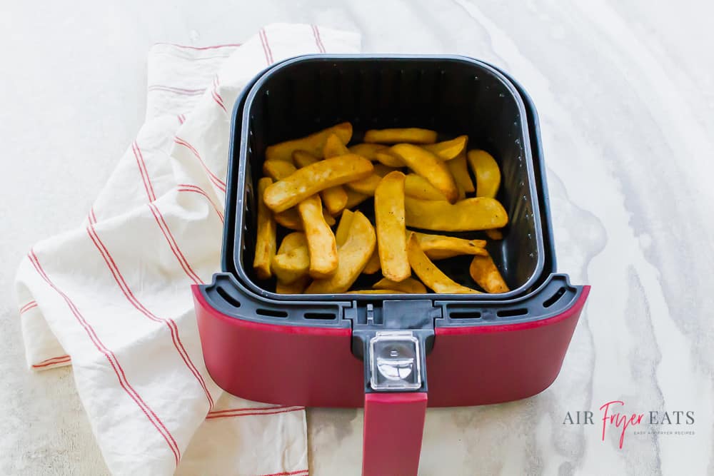 Horizontal picture with steak fries in a red and black air fryer basket set on top of a white background. There is a white napkin with thin red stripes to the left of the basket.