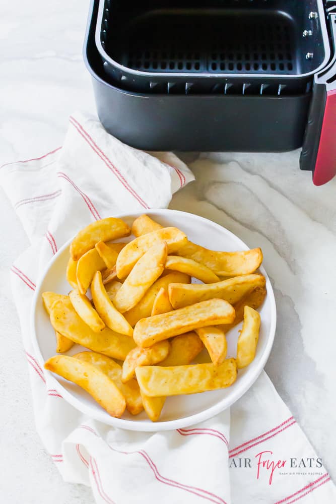 Vertical photo of steak fries on a white plate on top of a white napkin with thin red stripes. A red and black empty air fryer basket in the top right of picture.