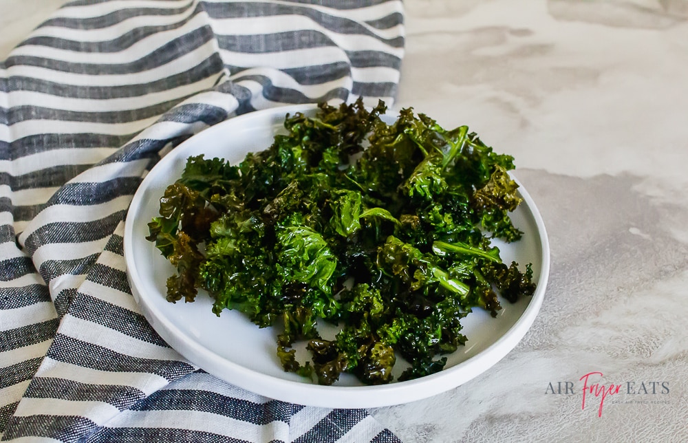 kale chips on a white plate with a blue striped kitchen towel on a marble countertop