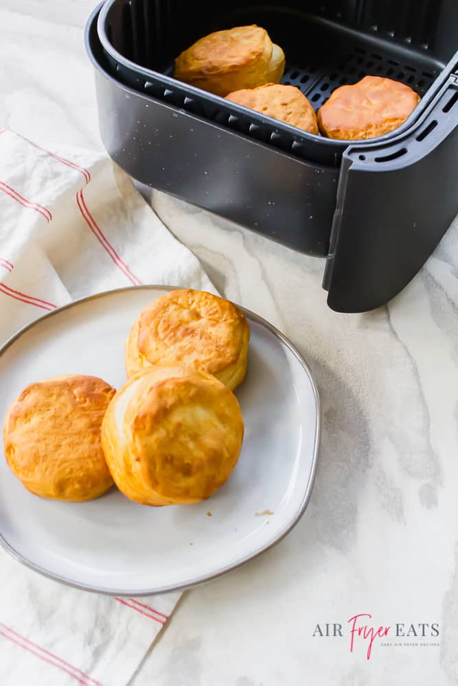 Vertical Image of air fryer biscuits on a white plate at the bottom of the picture and in an air fryer basket at the top of the photo.