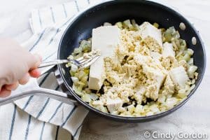 tofu being mashed with a fork in a skillet of onions and oil