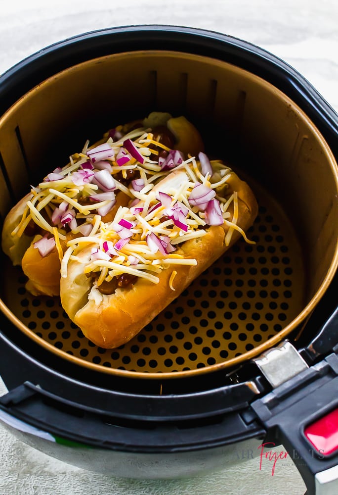 chili cheese dogs topped with diced red onion in an air fryer basket