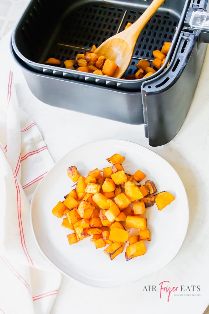 Image of cooked butternut squash cubes on a white plate and also in a black air fryer basket with a wooden spoon