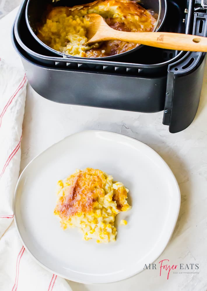 overhead shot of corn pudding on white plate and a bowl of creamed corn casserole in a black air fryer basket with a wooden spoon